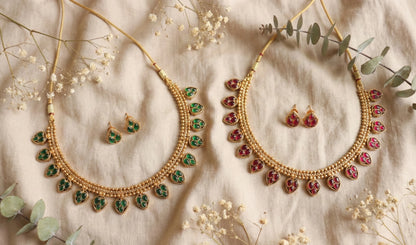 Gold toned necklaces with green and red gemstones on a neutral background with flowers and leaves.