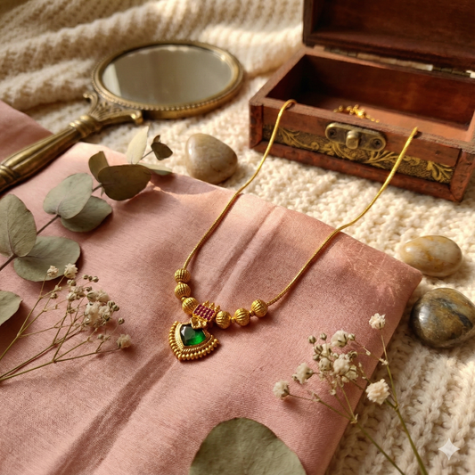 Gold plated necklace with a green palakka on a pink fabric with a wooden box and mirror in the background.