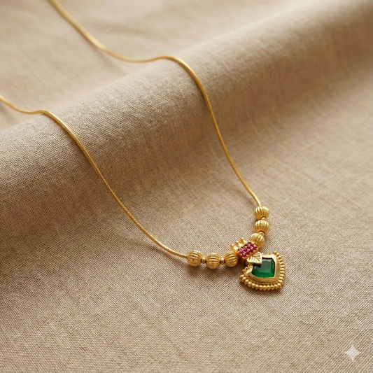 Gold plated necklace with a heart-shaped palakka pendant featuring green and red stones on a beige fabric background